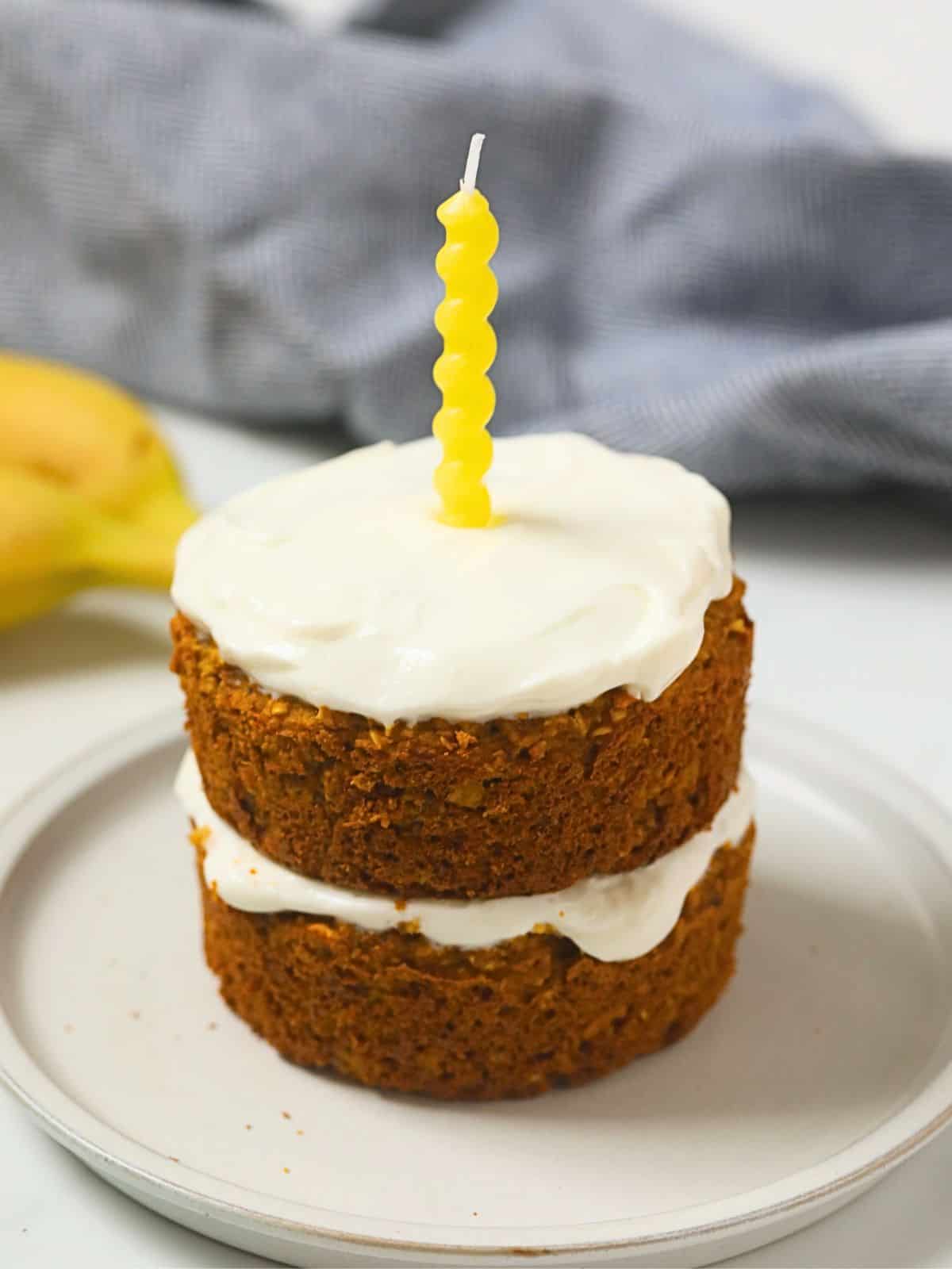 Two-layer dog birthday cake with oats, peanut butter, banana, and pumpkin, topped with Greek yogurt frosting with yellow candle.