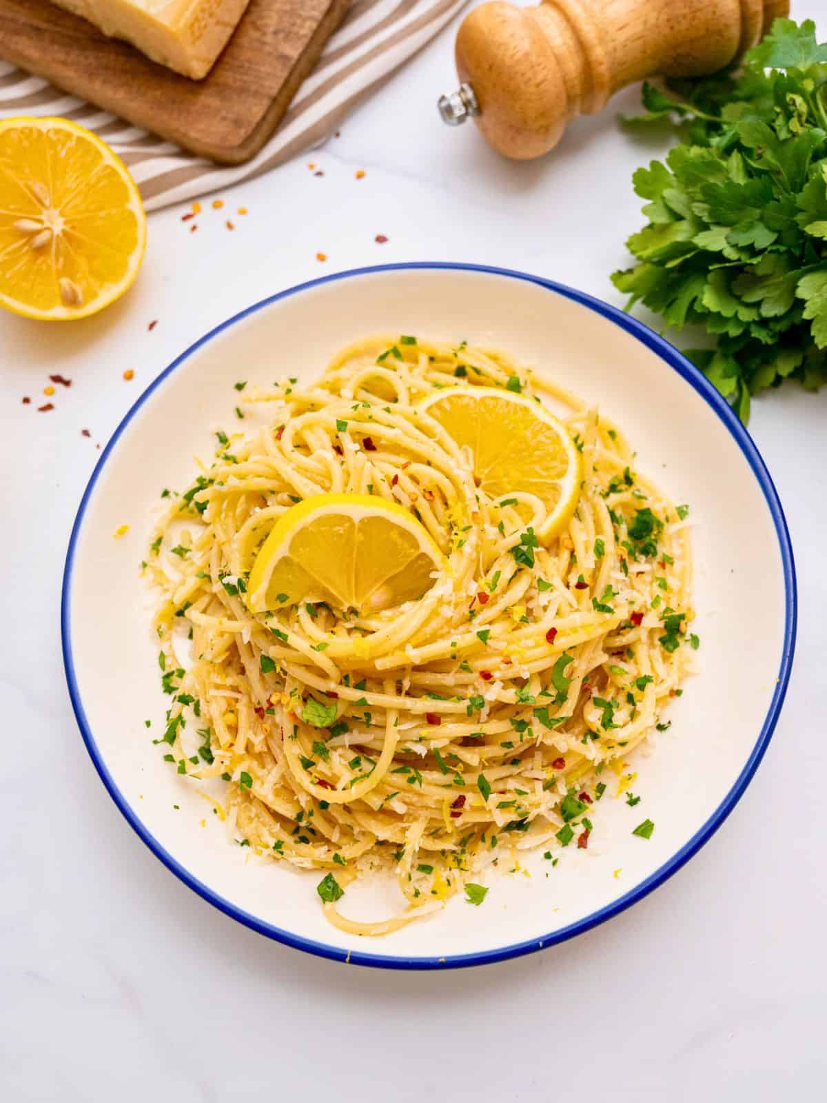 Spaghetti tossed with lemon, garlic butter, and parmesan in white bowl topped with lemon slices and fresh minced parsley.
