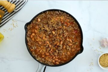 Deep skillet with ground beef, pinto beans, uncooked rice, beef stock, and salsa before baking.