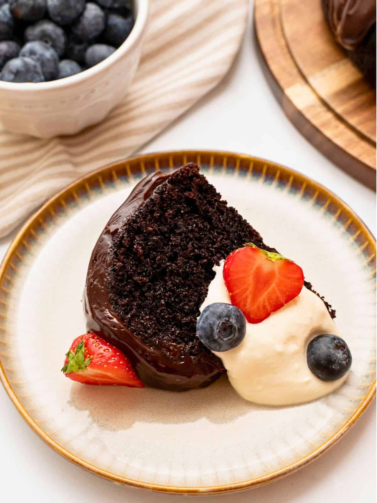 Slice of sour cream chocolate bundt cake iced with ganache topping on plate with berries and whipped cream.