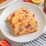 Slices of strawberry bread with lemon glaze on white plate board next to fresh lemons and fresh strawberries.