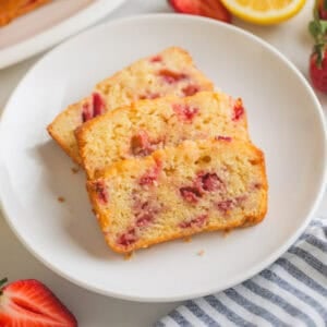 Slices of strawberry bread with lemon glaze on white plate board next to fresh lemons and fresh strawberries.