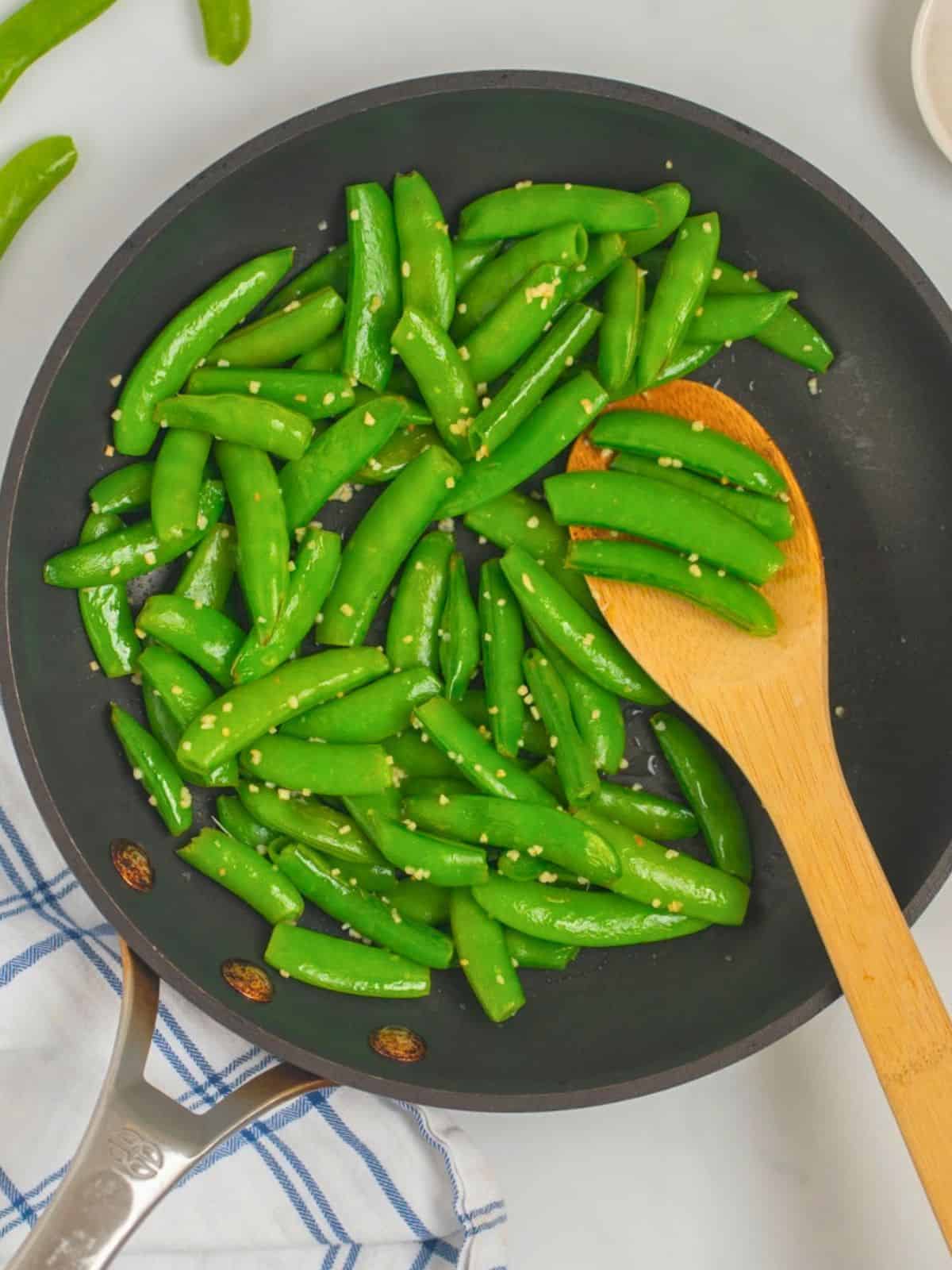 Sugar Snap Peas Wooden spoon scooping out sauteed sugar snap peas from skillet that have been lightly steamed and tossed with garlic and butter.