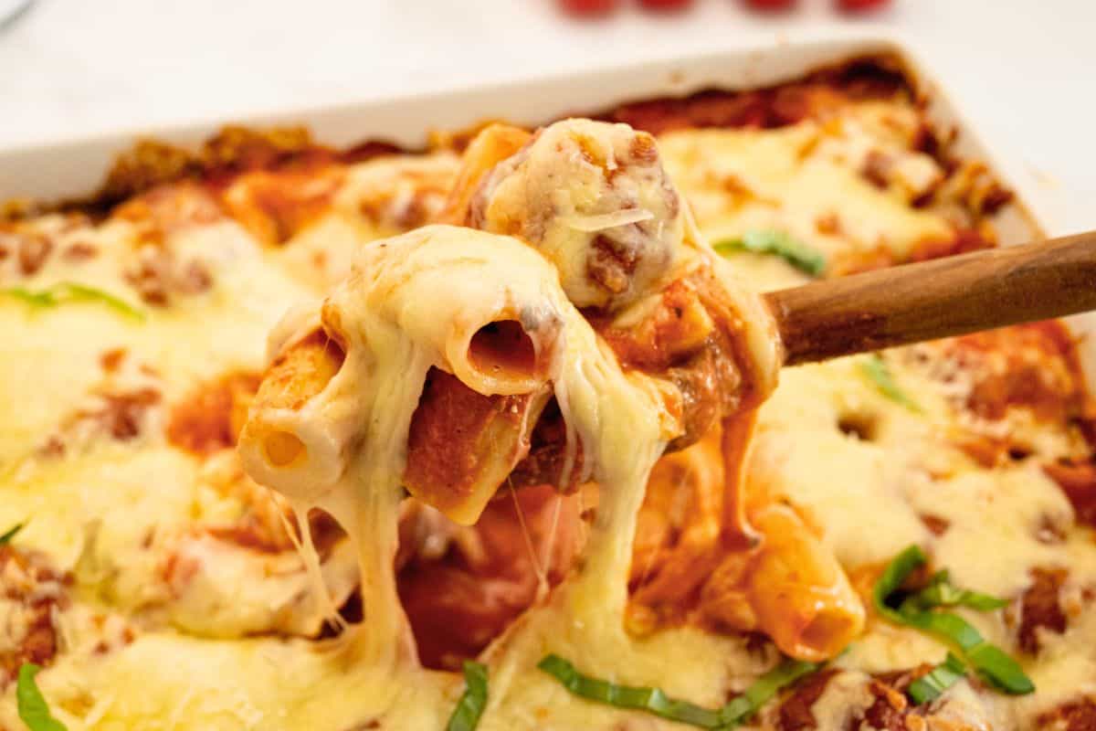 Spoonful of tender rigatoni noodles covered in creamy red sauce and melted mozzarella coming from large baking dish.