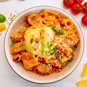 Bowl of cheesy baked pasta with creamy red sauce and sliced sausage topped with ribbons of basil.