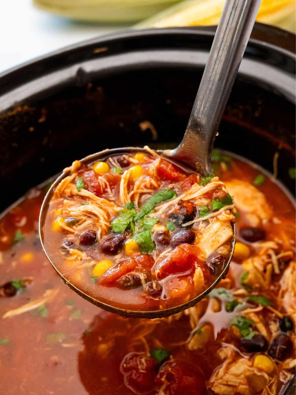 Ladle dishing out scoop of chicken tortilla soup from slow cooker with black beans, corn, and shredded chicken.