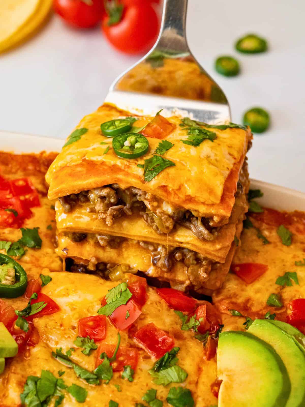 Spatula pulling out slice of baked beef enchilada casserole from casserole pan showing layers of ground beef, cheese, tortillas, and enchilada sauce. St