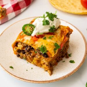 Slice of stacked beef enchiladas showing layers of seasoned ground beef, black beans, cheese, tortillas, and enchilada sauce topped with sour cream and cilantro.