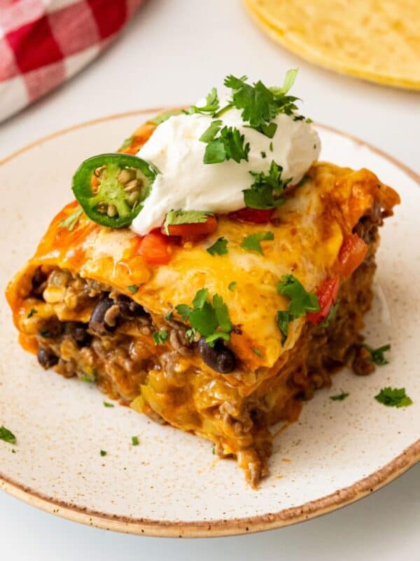 Slice of stacked beef enchiladas showing layers of seasoned ground beef, black beans, cheese, tortillas, and enchilada sauce topped with sour cream and cilantro.