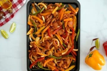 Sliced chicken breast, raw bell peppers, and yellow onion on sheet pan tossed with fajita seasoning.