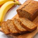 Sliced whole wheat banana bread on wooden cutting board with fresh bananas in background.