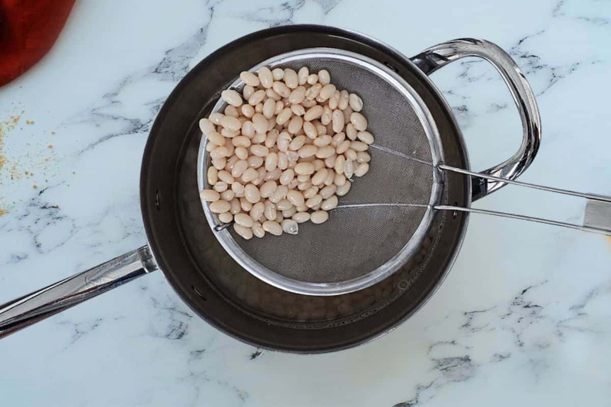 Spoonful of cooked navy beans in spider spoon over saucepan.