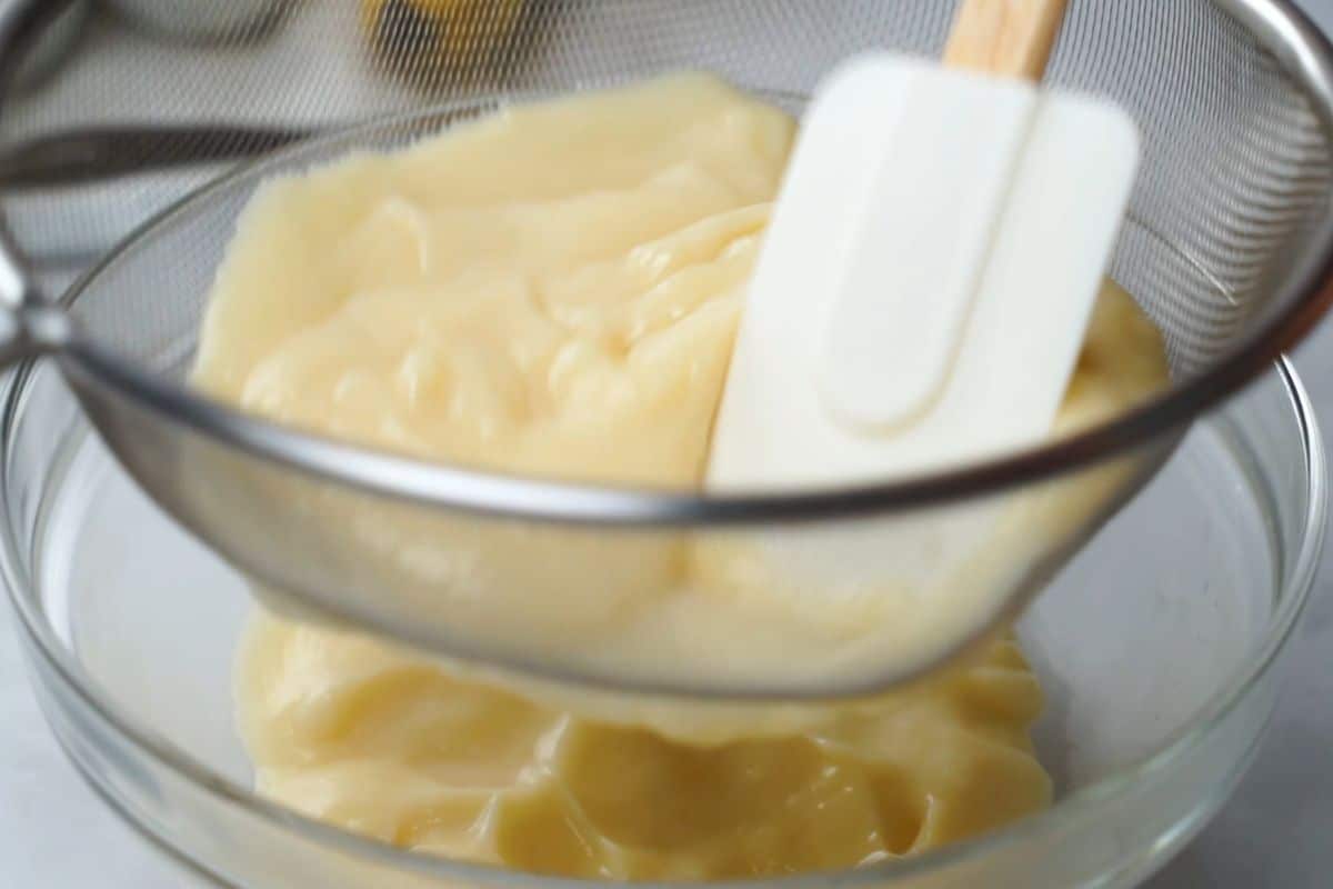 Homemade vanilla pudding being pushed through fine mesh strainer with rubber spatula.