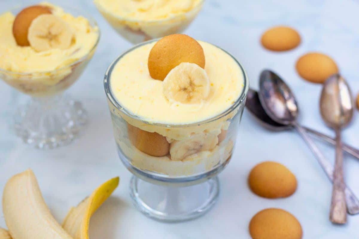 Homemade vanilla pudding layered with sliced bananas and vanilla wafters in mini trifle dishes next to fresh bananas and vanilla wafers.