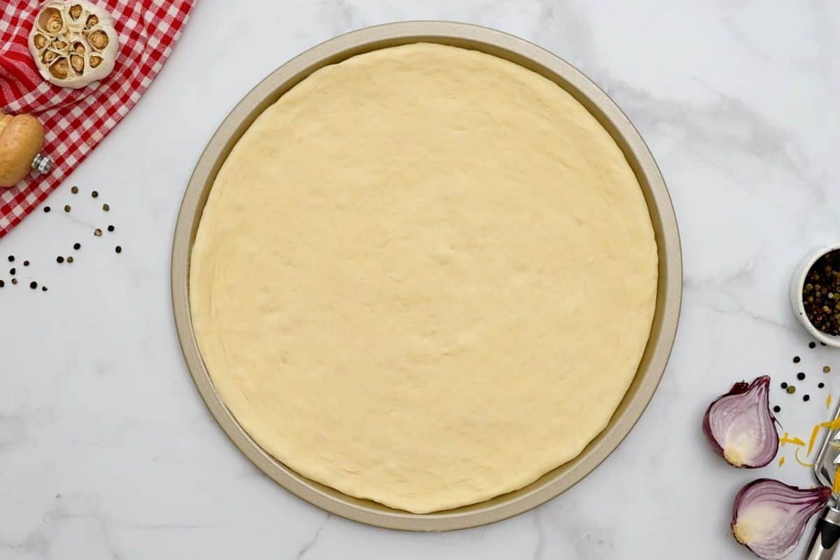 BBQ Chicken Pizza Recipe Pizza dough stretched out on a 12-inch pizza pan next to BBQ sauce and red onions.