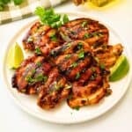 Grilled cilantro lime chicken thighs on platter topped with fresh cilantro next to fresh lime wedges.