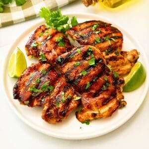 Grilled cilantro lime chicken thighs on platter topped with fresh cilantro next to fresh lime wedges.