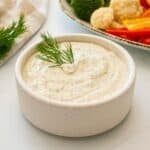 Homemade dill dip made with dried dill weed in white bowl topped with fresh dill and fresh veggies in background.