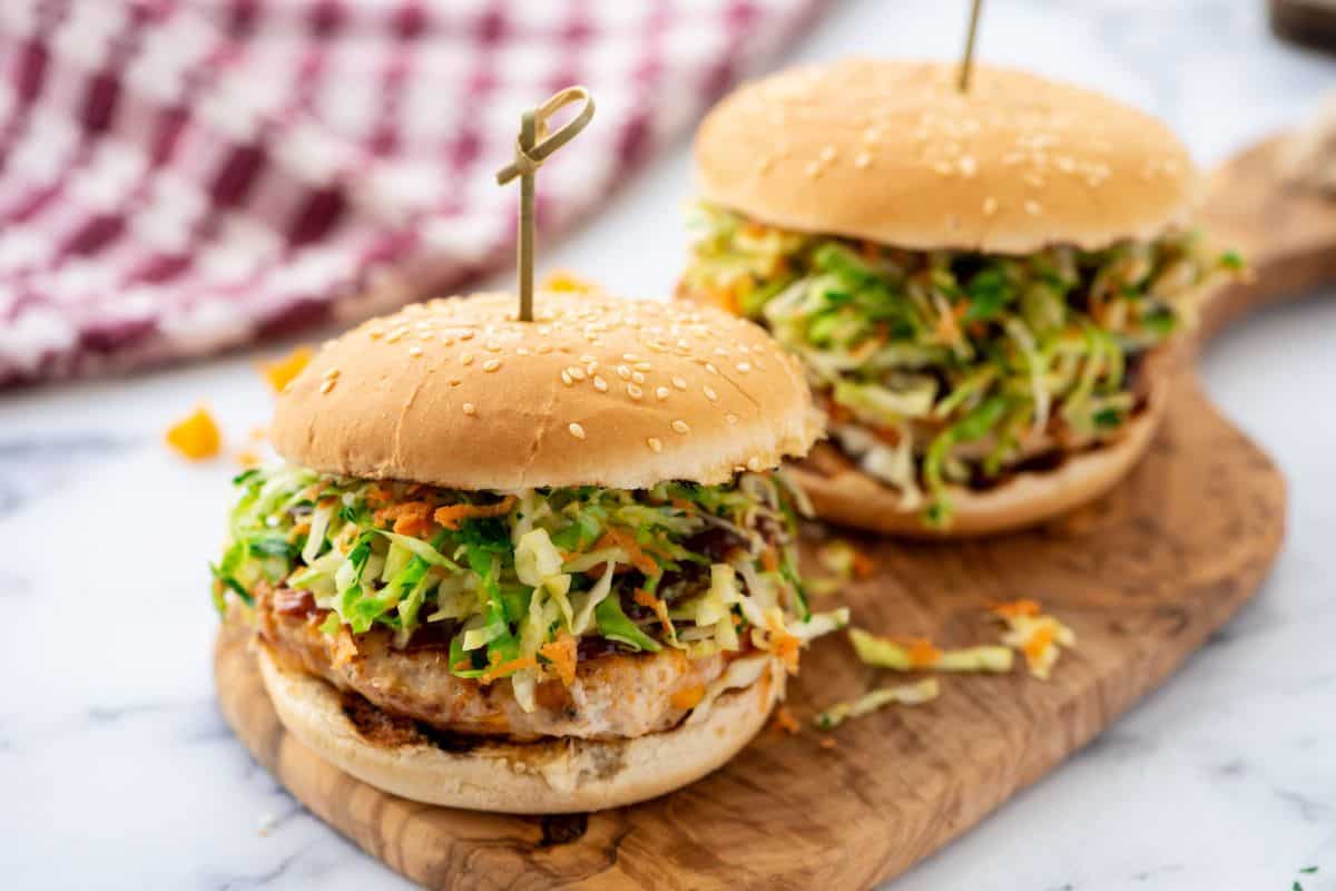 Two grilled BBQ chicken burgers made with BBQ sauce and cheese on platter topped homemade coleslaw served on sesame buns.