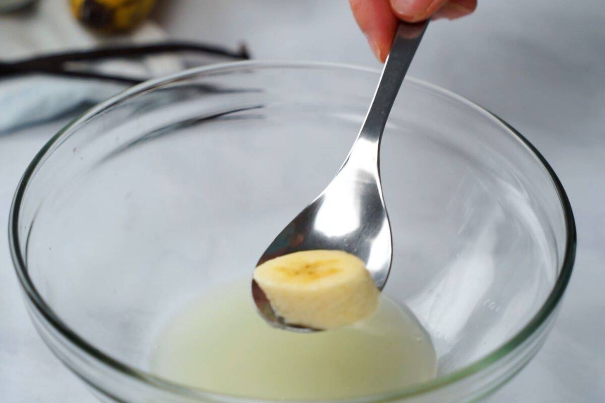 Spoon with sliced banana being dipped into lemon juice to prevent browning in banana pudding recipe.