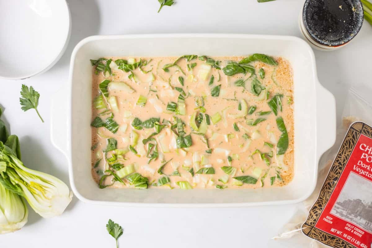 Sliced bok choy mixed into coconut milk, soy sauce, sesame oil, curry paste, and rice vinegar in 9x13 inch baking dish next to bag of frozen chicken and pork potstickers.