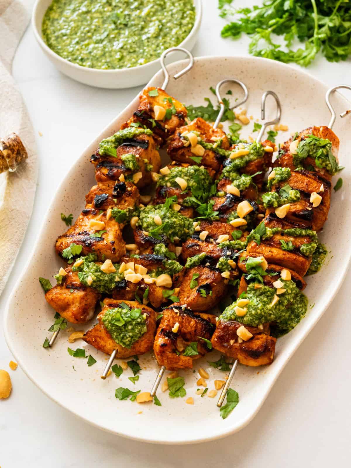 Grilled chicken kabobs on white platter topped with cilantro pesto, fresh cilantro, and chopped peanuts.