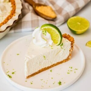 Slice of no bake key lime pie with graham cracker crust on plate topped with piped whipped cream, a lime slice, and lime zest.