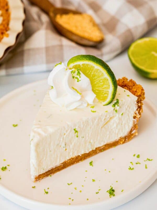 Slice of no bake key lime pie with graham cracker crust on plate topped with piped whipped cream, a lime slice, and lime zest.