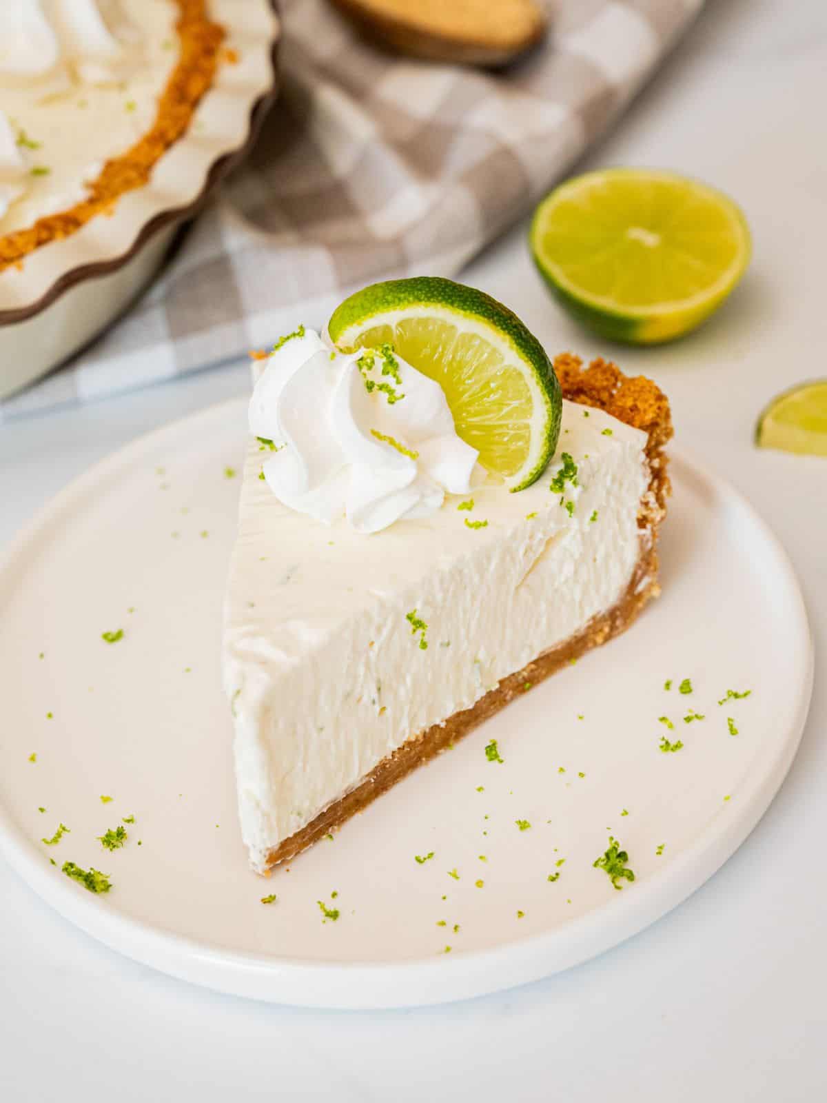 Easy Key Lime Pie Recipe Slice of homemade key lime pie with graham cracker crust on plate topped with piped whipped cream, a lime slice, and lime zest.