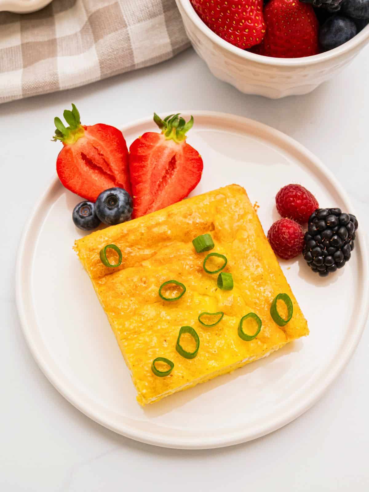 Slice of cottage cheese egg bake on white plate topped with sliced green onions and berries on the side.