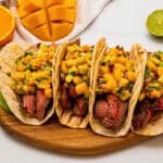 4 soft tortillas filled with sliced grilled marinated flank steak and topped with mango avocado salsa next to fresh mangoes, limes, and oranges.