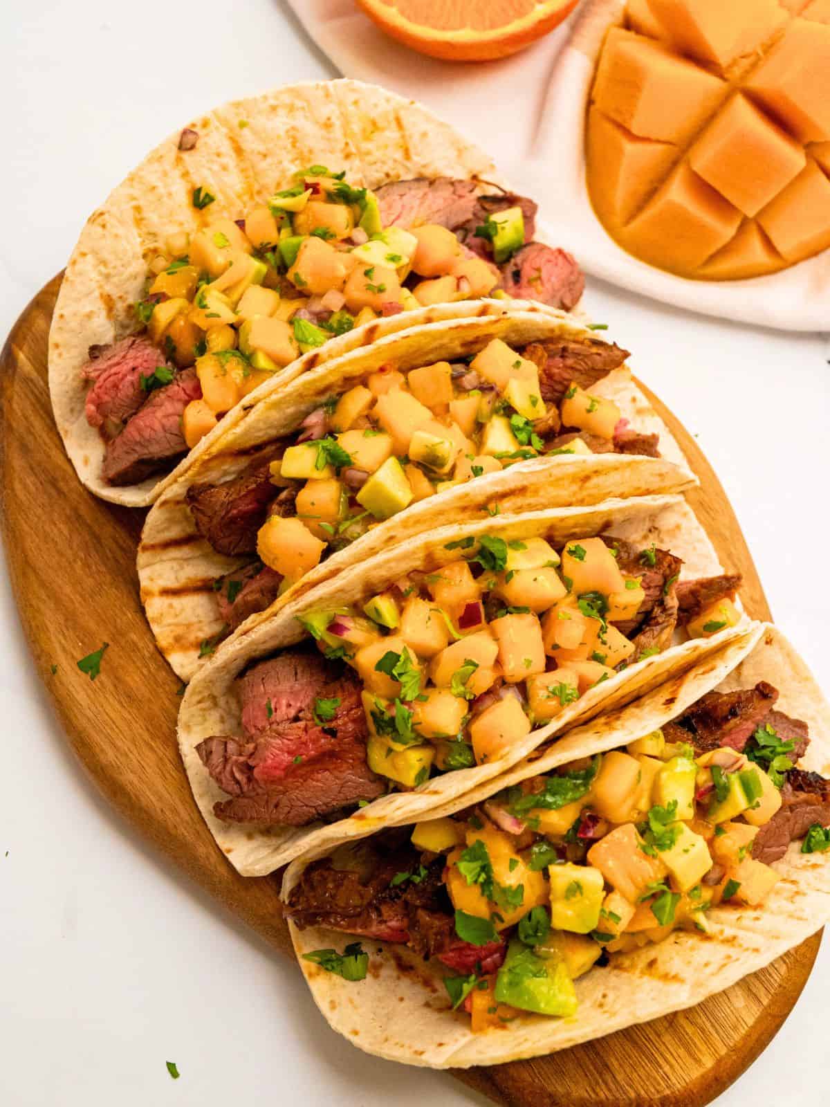 4 soft tortillas filled with sliced grilled marinated flank steak and topped with mango avocado salsa next to fresh mango.