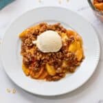 Homemade peach crisp with oat topping and sliced fresh peaches served with scoop of vanilla ice cream.