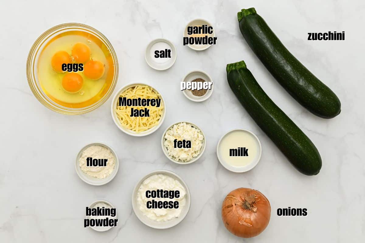 Savory Zucchini Pie Recipe Zucchini, yellow onion, cottage cheese, feta, Monterey Jack cheese, milk, eggs, seasonings, and baking mix labeled on counter.