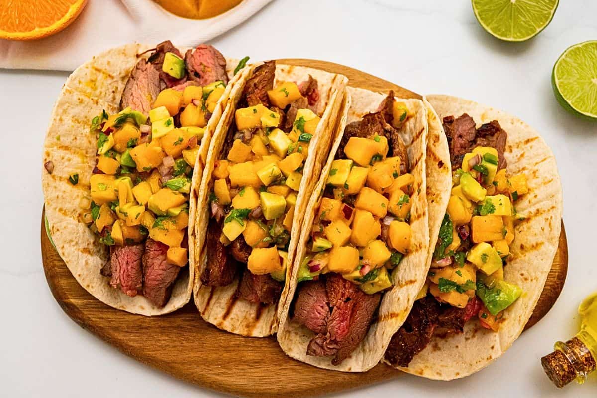 4 soft tortillas filled with sliced grilled marinated flank steak and topped with mango avocado salsa next to fresh mango.