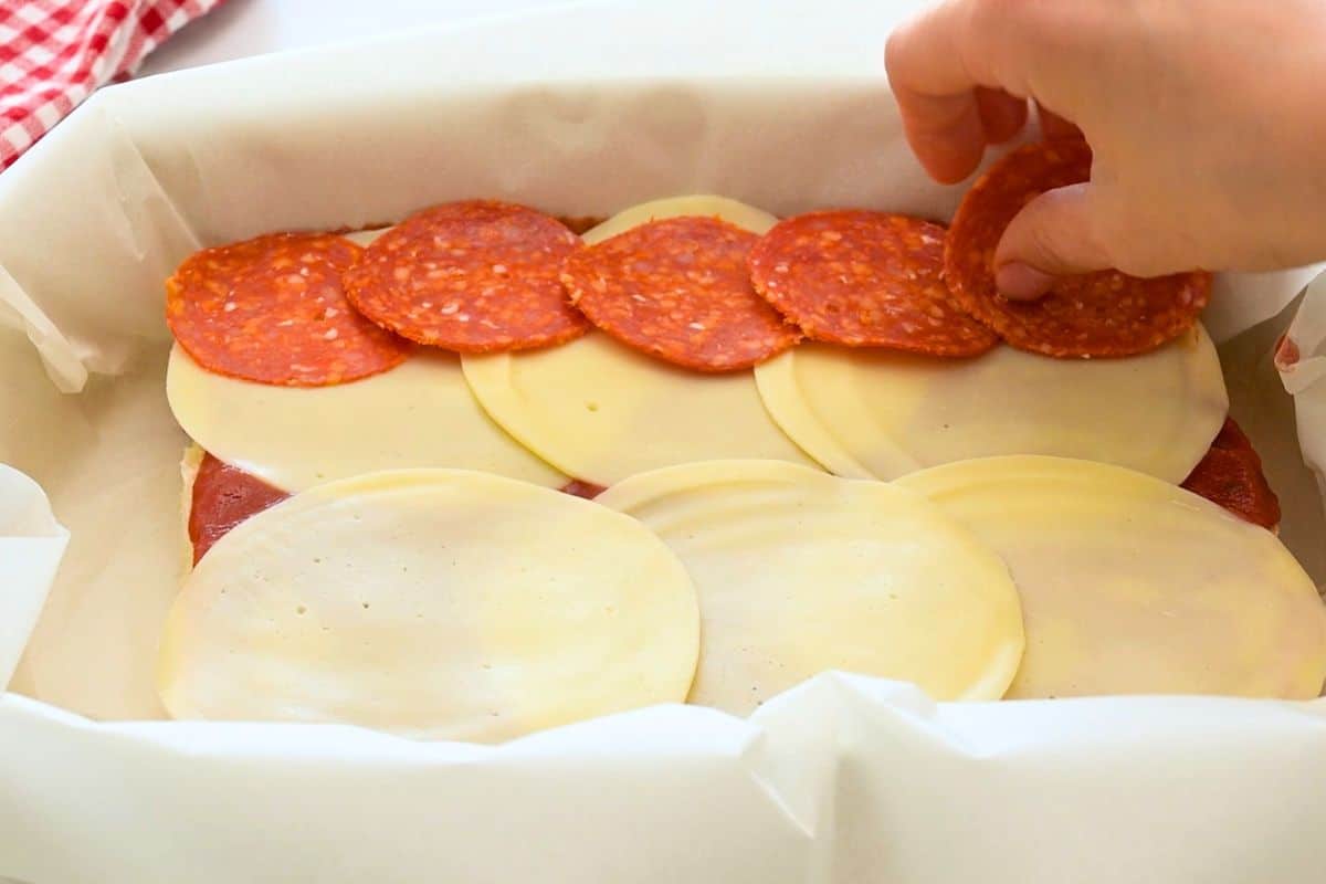 Layering pepperoni over bottom of slider rolls that have been topped with thin layer of pizza sauce and sliced provolone cheese.