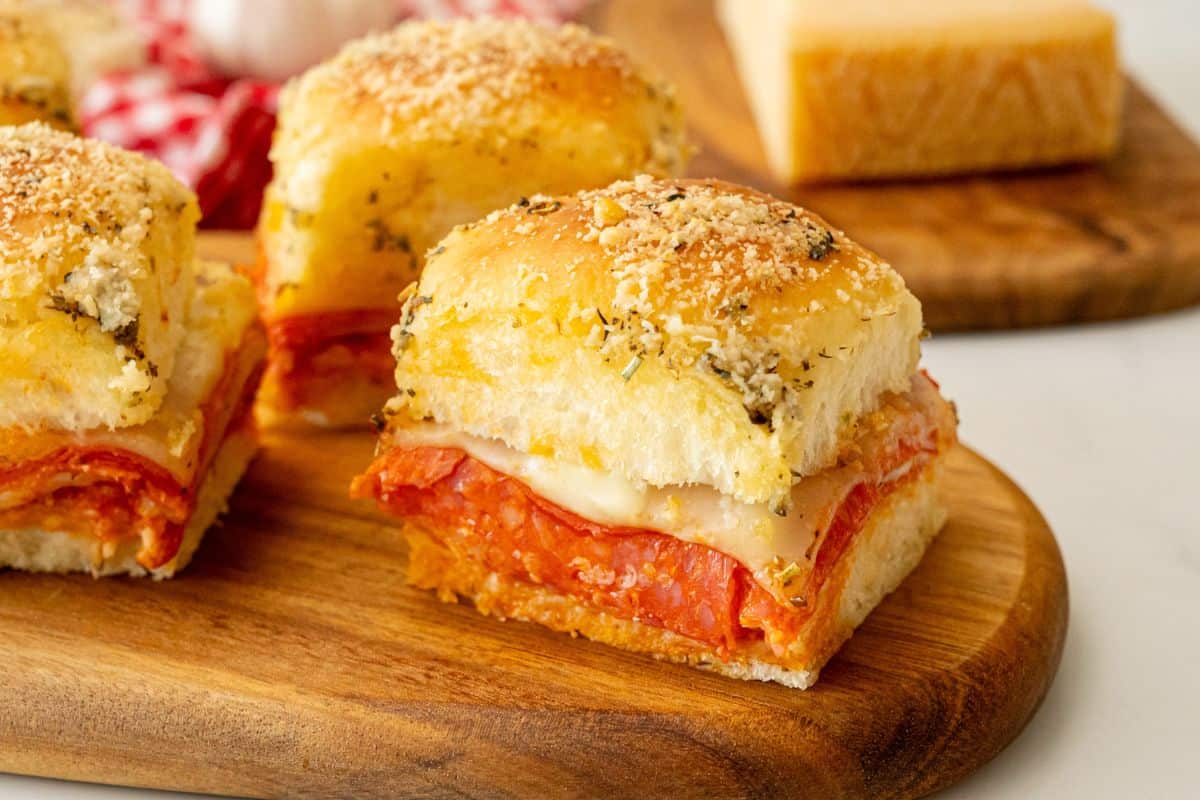 Sliced pizza sliders on wooden cutting board showing layers of provolone, pepperoni, pizza sauce, and mozzarella cheese on garlic butter slider bun.