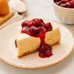 Slice of light and airy cheesecake with sour cream topping on plate topped with cherries.