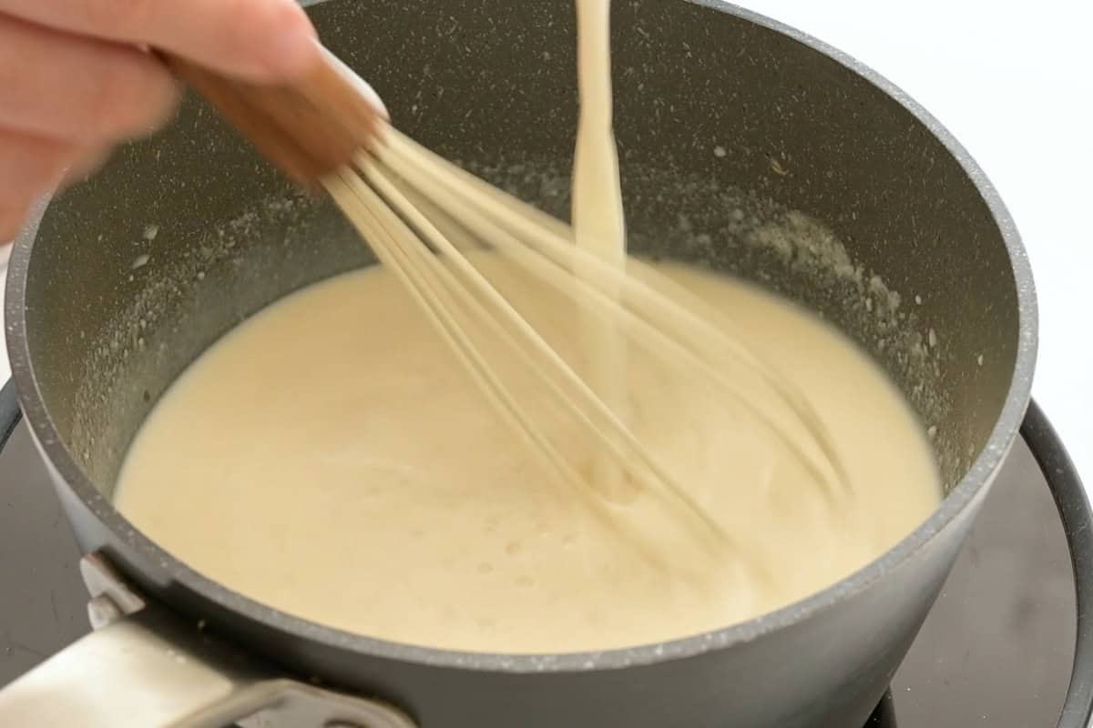 Velvety Baked Mac and Cheese Milk being drizzled into saucepan as it is being whisked into the roux.